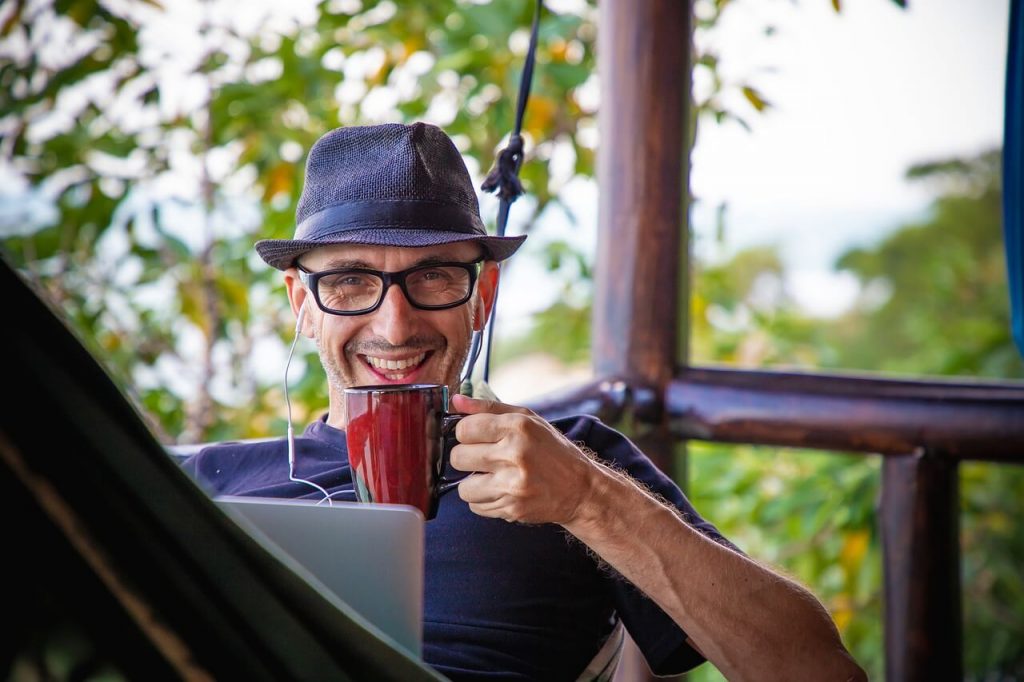 A man with a hat drinking and working on a laptop